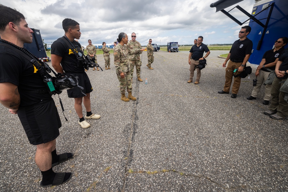 A military briefing or coordination meeting is taking place on what appears to be a tarmac or airfield. A soldier in camouflage uniform appears to be addressing a mixed group of military personnel and civilians in black tactical uniforms. The setting suggests a joint operation or emergency response coordination, possibly related to disaster relief or a military exercise.