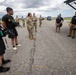 A military briefing or coordination meeting is taking place on what appears to be a tarmac or airfield. A soldier in camouflage uniform appears to be addressing a mixed group of military personnel and civilians in black tactical uniforms. The setting suggests a joint operation or emergency response coordination, possibly related to disaster relief or a military exercise.