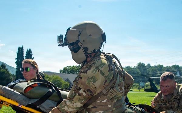 U.S. Army personnel and a civilian medical worker are transferring a patient on a Stryker stretcher from a helicopter landing zone to an ambulance. At least three individuals are actively handling the gurney, with a military helicopter (likely a UH-60 Black Hawk) visible in the background. This appears to be either a medical evacuation (MEDEVAC) operation or a training exercise.