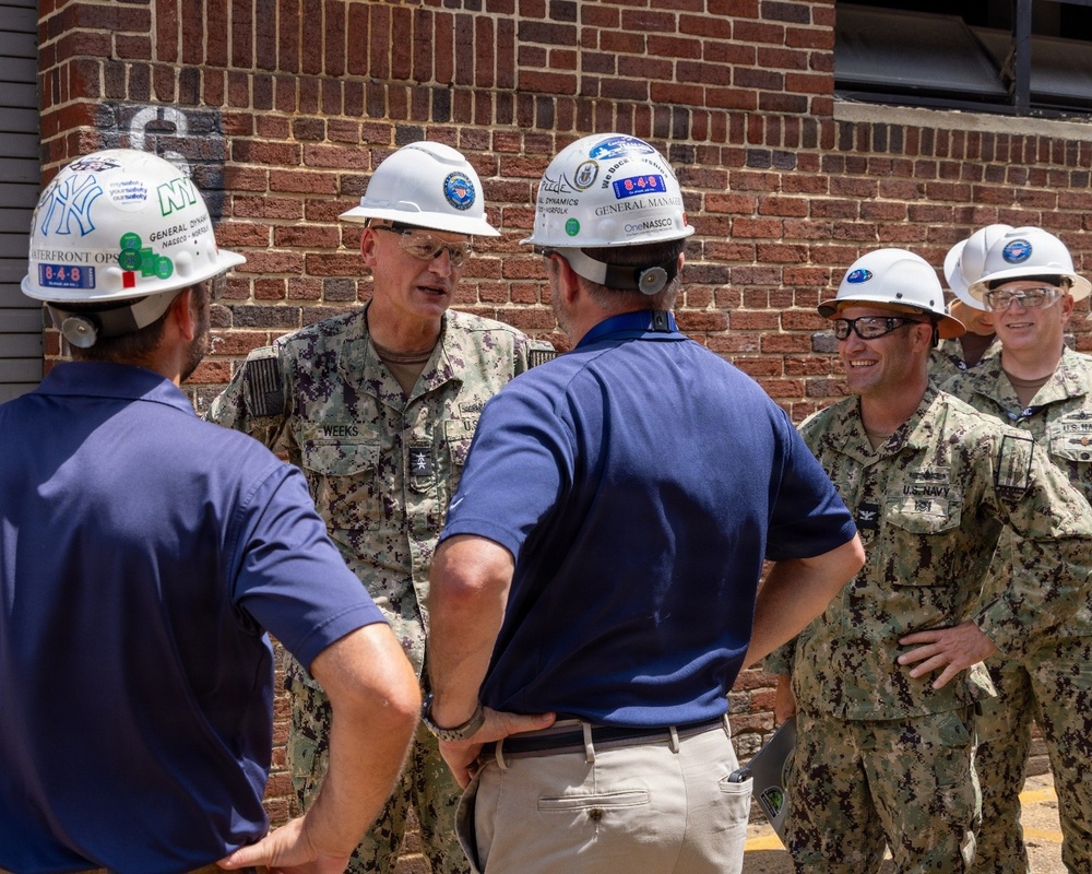 A group of U.S. Navy personnel in digital camouflage uniforms are engaged in conversation with civilian contractors wearing white hard hats outside a brick building. The naval officer in the center bears the name tape 'WEEKS' and appears to be of senior rank based on visible insignia. The hard hats display 'General Dynamics,' 'NASCO,' and 'OneNASCO' branding, suggesting this is a naval shipyard or facility inspection.