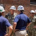 A group of U.S. Navy personnel in digital camouflage uniforms are engaged in conversation with civilian contractors wearing white hard hats outside a brick building. The naval officer in the center bears the name tape 'WEEKS' and appears to be of senior rank based on visible insignia. The hard hats display 'General Dynamics,' 'NASCO,' and 'OneNASCO' branding, suggesting this is a naval shipyard or facility inspection.