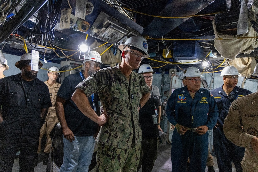 A group of U.S. Navy personnel and civilian workers are conducting an inspection or walkthrough in a confined, industrial space aboard what appears to be a naval vessel. The central figure is a Navy officer in camouflage utilities wearing a white hard hat with safety glasses and ear protection. A name tag reading 'WEEKS' is visible on the central officer, and another individual's uniform references 'USS Bataan' and the name 'Rod Jacoby'.