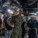 A group of U.S. Navy personnel and civilian workers are conducting an inspection or walkthrough in a confined, industrial space aboard what appears to be a naval vessel. The central figure is a Navy officer in camouflage utilities wearing a white hard hat with safety glasses and ear protection. A name tag reading 'WEEKS' is visible on the central officer, and another individual's uniform references 'USS Bataan' and the name 'Rod Jacoby'.