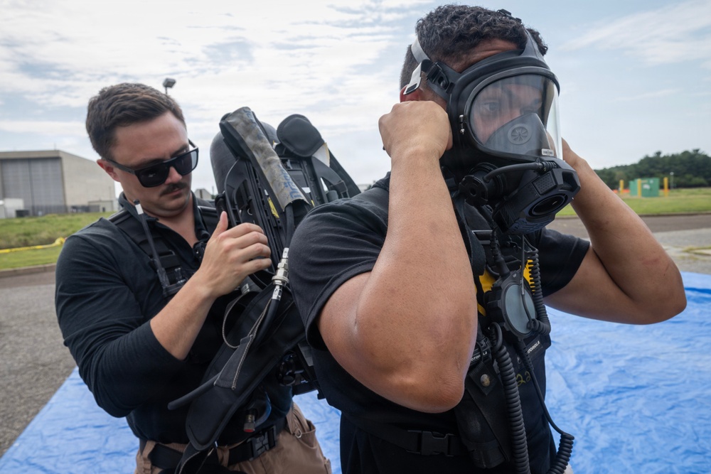 Two individuals dressed in black tactical clothing are engaged in what appears to be a hazmat or CBRN (Chemical, Biological, Radiological, Nuclear) training exercise. One person is donning a full-face respirator/gas mask while the other assists with securing a breathing apparatus or backpack unit. The setting appears to be an outdoor tarmac or industrial area, suggesting a first responder or military training environment.