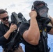 Two individuals dressed in black tactical clothing are engaged in what appears to be a hazmat or CBRN (Chemical, Biological, Radiological, Nuclear) training exercise. One person is donning a full-face respirator/gas mask while the other assists with securing a breathing apparatus or backpack unit. The setting appears to be an outdoor tarmac or industrial area, suggesting a first responder or military training environment.