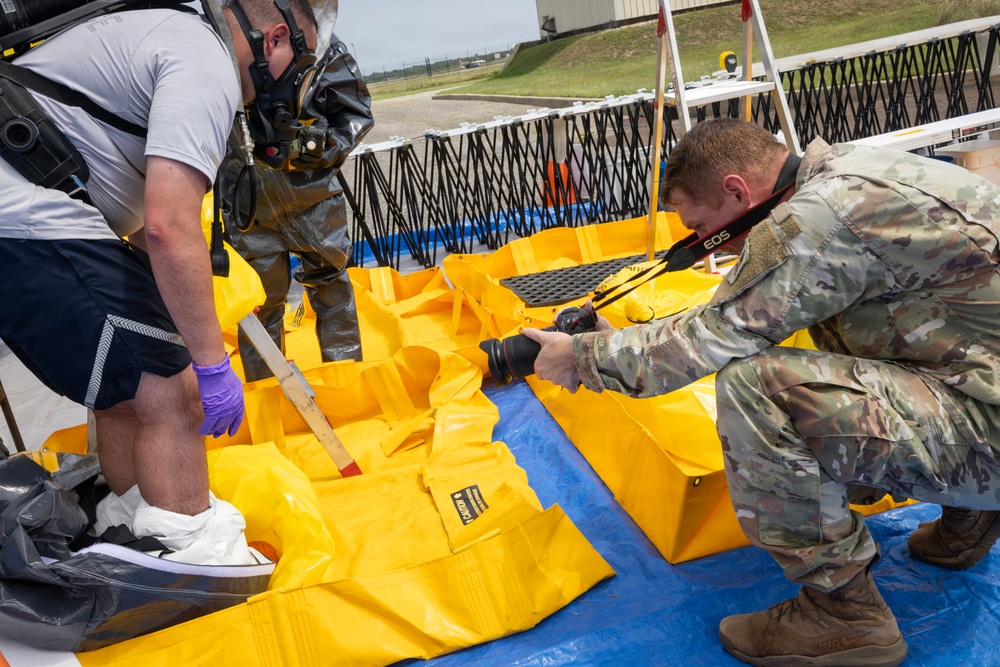 The image shows what appears to be a hazmat or emergency response training exercise at a military or industrial facility. Personnel in protective gear and military uniform are working with yellow containment equipment, while a soldier with a Canon EOS camera documents the activity. The scene includes spill containment berms, protective suits, and respiratory equipment consistent with CBRN or hazmat operations.