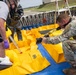 The image shows what appears to be a hazmat or emergency response training exercise at a military or industrial facility. Personnel in protective gear and military uniform are working with yellow containment equipment, while a soldier with a Canon EOS camera documents the activity. The scene includes spill containment berms, protective suits, and respiratory equipment consistent with CBRN or hazmat operations.
