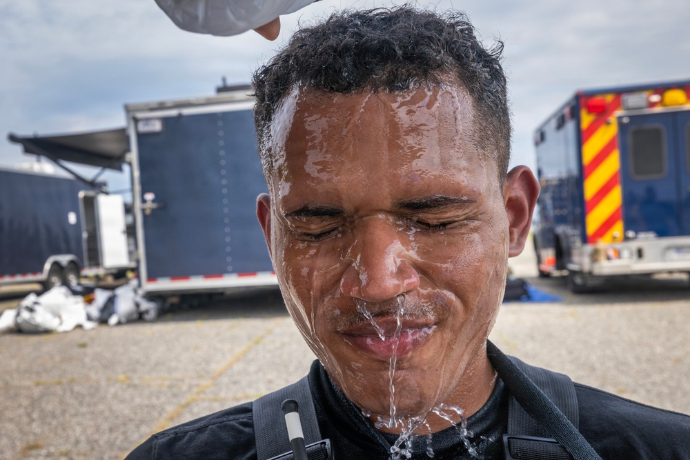 A man in a black tactical or uniform top is having water poured over his face and head by another person whose hand is partially visible at the top of the frame. His eyes are closed and water is streaming down his face. The setting appears to be an outdoor staging or operational area with emergency or utility vehicles visible in the background.