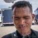 A man in a black tactical or uniform top is having water poured over his face and head by another person whose hand is partially visible at the top of the frame. His eyes are closed and water is streaming down his face. The setting appears to be an outdoor staging or operational area with emergency or utility vehicles visible in the background.