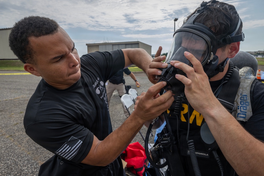 Two individuals are shown in an outdoor setting where one person is assisting the other in fitting or adjusting a full-face respirator/gas mask. The person wearing the mask has 'ARMY' visible on their clothing and is equipped with a Dräger breathing apparatus. This appears to be a military or emergency response training exercise.