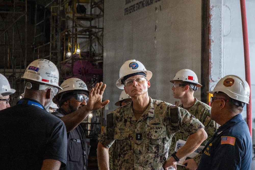 A group of individuals wearing white hard hats and safety glasses are gathered in what appears to be a large industrial or shipyard facility. A U.S. Navy officer in camouflage uniform with the name tape 'WEEKS' is centrally positioned, appearing to receive a briefing or tour. The background shows scaffolding, heavy machinery, and a large industrial structure.