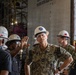 A group of individuals wearing white hard hats and safety glasses are gathered in what appears to be a large industrial or shipyard facility. A U.S. Navy officer in camouflage uniform with the name tape 'WEEKS' is centrally positioned, appearing to receive a briefing or tour. The background shows scaffolding, heavy machinery, and a large industrial structure.