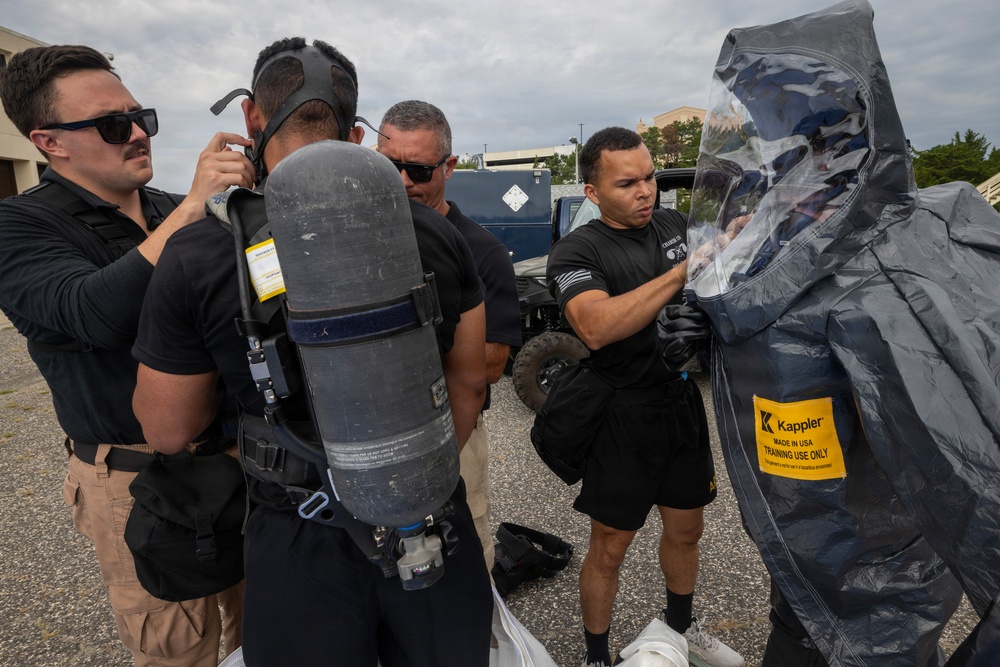A group of personnel in black uniforms are conducting a hazmat or CBRN training exercise outdoors. One individual is being suited up in a Kappler protective encapsulating suit labeled 'Training Use Only,' while another person has an SCBA (Self-Contained Breathing Apparatus) tank on their back. Multiple team members are assisting with donning the protective equipment.