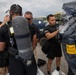 A group of personnel in black uniforms are conducting a hazmat or CBRN training exercise outdoors. One individual is being suited up in a Kappler protective encapsulating suit labeled 'Training Use Only,' while another person has an SCBA (Self-Contained Breathing Apparatus) tank on their back. Multiple team members are assisting with donning the protective equipment.