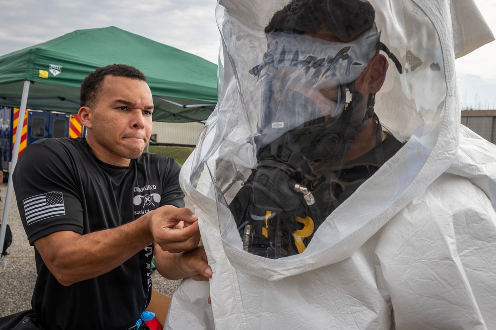 A person in a black t-shirt with a US flag patch and text reading 'CBRLIECO 84th Chem' is assisting another individual in donning or adjusting a white hazmat/CBRN protective suit with a clear face shield. The setting appears to be an outdoor training or exercise area with a green canopy tent and what appears to be an emergency vehicle in the background. The activity suggests a chemical, biological, radiological, or nuclear (CBRN) training exercise.