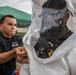 A person in a black t-shirt with a US flag patch and text reading 'CBRLIECO 84th Chem' is assisting another individual in donning or adjusting a white hazmat/CBRN protective suit with a clear face shield. The setting appears to be an outdoor training or exercise area with a green canopy tent and what appears to be an emergency vehicle in the background. The activity suggests a chemical, biological, radiological, or nuclear (CBRN) training exercise.