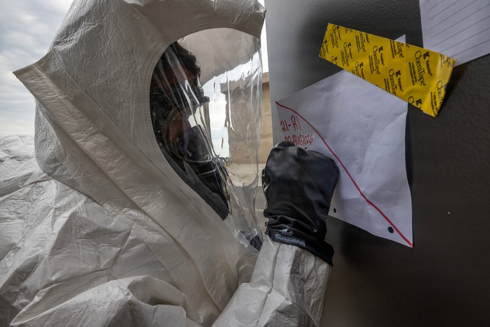 A person wearing a white hazmat/CBRN protective suit with a full-face respirator mask and black gloves is writing or pointing at a paper document affixed to a surface. The document appears to have handwritten red text and is secured with yellow caution/chemical tape. The setting appears to be an outdoor or semi-outdoor environment during what looks like a hazmat exercise or response operation.
