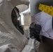 A person wearing a white hazmat/CBRN protective suit with a full-face respirator mask and black gloves is writing or pointing at a paper document affixed to a surface. The document appears to have handwritten red text and is secured with yellow caution/chemical tape. The setting appears to be an outdoor or semi-outdoor environment during what looks like a hazmat exercise or response operation.