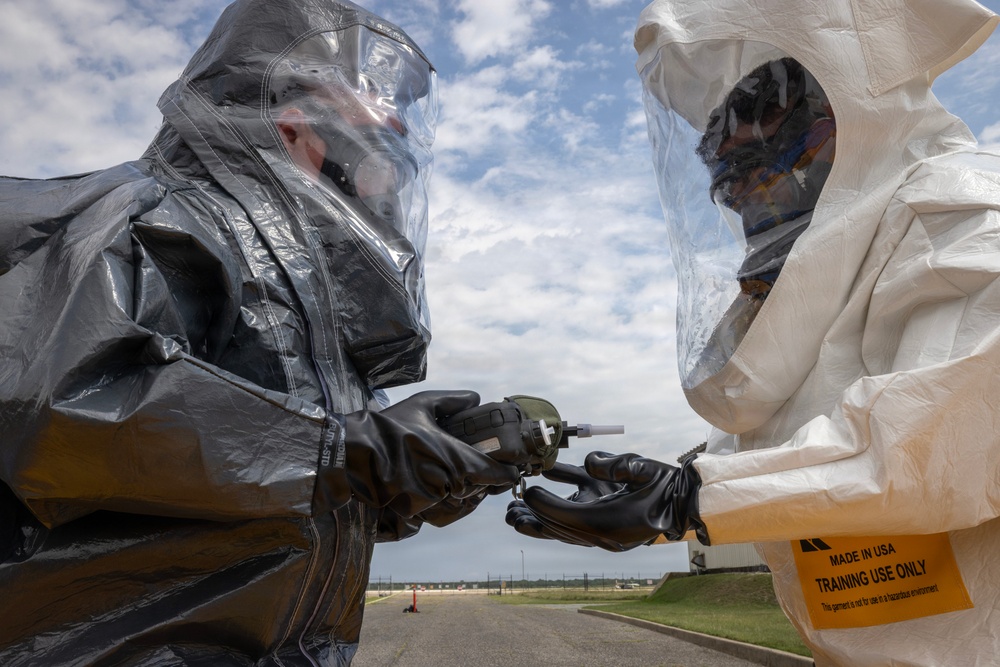 Two individuals wearing full hazmat/CBRN protective suits are engaged in a training exercise, handling what appears to be a detection or sampling device. The person on the right is wearing a white suit with a visible label reading 'MADE IN USA - TRAINING USE ONLY,' confirming this is a simulated exercise rather than a real hazardous environment response. The setting appears to be an outdoor military or emergency response training facility.