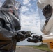 Two individuals wearing full hazmat/CBRN protective suits are engaged in a training exercise, handling what appears to be a detection or sampling device. The person on the right is wearing a white suit with a visible label reading 'MADE IN USA - TRAINING USE ONLY,' confirming this is a simulated exercise rather than a real hazardous environment response. The setting appears to be an outdoor military or emergency response training facility.