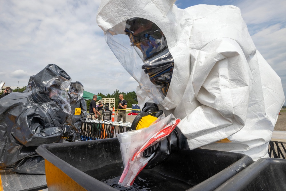 Personnel in full hazmat/CBRN protective suits are conducting what appears to be a decontamination exercise or operation. One person in a white Tyvek-style encapsulated suit and another in a black hazmat suit are working over a large black containment tub filled with water. Additional personnel and equipment are visible in the background, suggesting an organized training or response scenario.