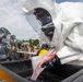 Personnel in full hazmat/CBRN protective suits are conducting what appears to be a decontamination exercise or operation. One person in a white Tyvek-style encapsulated suit and another in a black hazmat suit are working over a large black containment tub filled with water. Additional personnel and equipment are visible in the background, suggesting an organized training or response scenario.