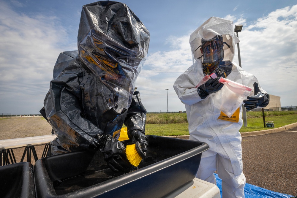 Two individuals wearing full hazmat/CBRN protective suits are engaged in what appears to be a decontamination training exercise outdoors. One person in a black encapsulating suit is scrubbing inside a large containment tub using a yellow brush, while a second person in a white Tyvek-style suit holds a clear evidence or sample bag. A yellow tag on the white suit reads 'TRAINING USE ONLY', confirming this is a drill or exercise.