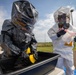 Two individuals wearing full hazmat/CBRN protective suits are engaged in what appears to be a decontamination training exercise outdoors. One person in a black encapsulating suit is scrubbing inside a large containment tub using a yellow brush, while a second person in a white Tyvek-style suit holds a clear evidence or sample bag. A yellow tag on the white suit reads 'TRAINING USE ONLY', confirming this is a drill or exercise.