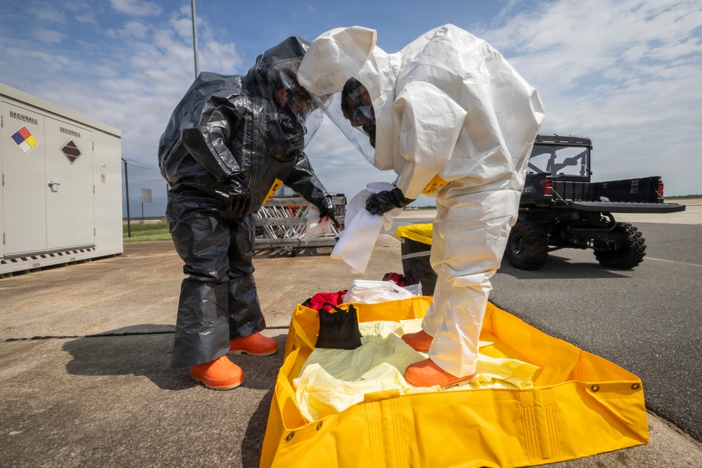 Two personnel in full hazardous materials (HAZMAT) protective suits are working together on a yellow containment berm or decontamination pad on a paved surface. One individual wears a dark/black encapsulated suit with orange boots, while the other wears a white Tyvek-style suit, both equipped with full-face respirators and black gloves. The setting appears to be an airfield or industrial facility, suggesting a HAZMAT training exercise or actual decontamination operation.