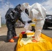 Two personnel in full hazardous materials (HAZMAT) protective suits are working together on a yellow containment berm or decontamination pad on a paved surface. One individual wears a dark/black encapsulated suit with orange boots, while the other wears a white Tyvek-style suit, both equipped with full-face respirators and black gloves. The setting appears to be an airfield or industrial facility, suggesting a HAZMAT training exercise or actual decontamination operation.