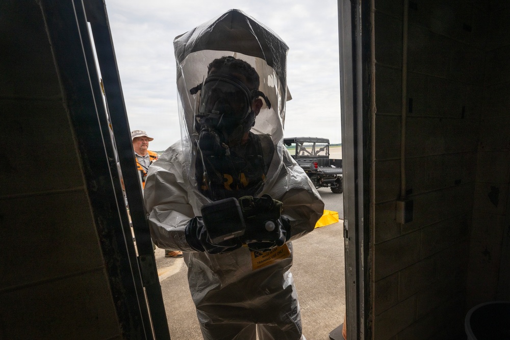A person wearing a full hazmat/CBRN protective suit with an integrated gas mask and clear hood is entering or exiting a doorway. The suit appears to be a Level B or Level C encapsulating protective ensemble, likely used in a training exercise based on a visible 'TRAINING' label on the suit. A second person in orange safety vest and hat is visible in the background near a utility vehicle.