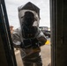 A person wearing a full hazmat/CBRN protective suit with an integrated gas mask and clear hood is entering or exiting a doorway. The suit appears to be a Level B or Level C encapsulating protective ensemble, likely used in a training exercise based on a visible 'TRAINING' label on the suit. A second person in orange safety vest and hat is visible in the background near a utility vehicle.