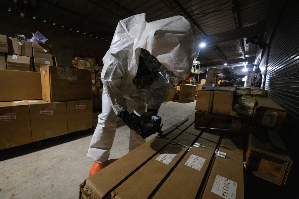 A person wearing a full white hazmat/protective suit with a respirator mask and orange gloves is using a handheld detection device to scan flat cardboard packages in a warehouse or storage facility. The warehouse contains numerous cardboard boxes, some labeled 'FRAGILE,' stacked throughout the space. Additional personnel are visible in the background near a roll-up door with artificial lighting.