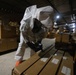 A person wearing a full white hazmat/protective suit with a respirator mask and orange gloves is using a handheld detection device to scan flat cardboard packages in a warehouse or storage facility. The warehouse contains numerous cardboard boxes, some labeled 'FRAGILE,' stacked throughout the space. Additional personnel are visible in the background near a roll-up door with artificial lighting.