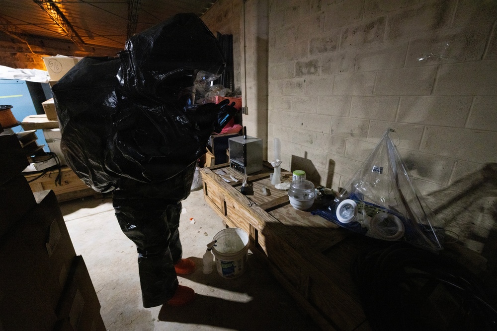A person wearing a full black hazmat/protective suit is examining a makeshift workbench in what appears to be a basement or warehouse space. The workbench, constructed from wooden pallets, holds various containers, glassware, and equipment. The setting has concrete block walls and appears to be a law enforcement or emergency response documentation scene.
