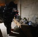 A person wearing a full black hazmat/protective suit is examining a makeshift workbench in what appears to be a basement or warehouse space. The workbench, constructed from wooden pallets, holds various containers, glassware, and equipment. The setting has concrete block walls and appears to be a law enforcement or emergency response documentation scene.
