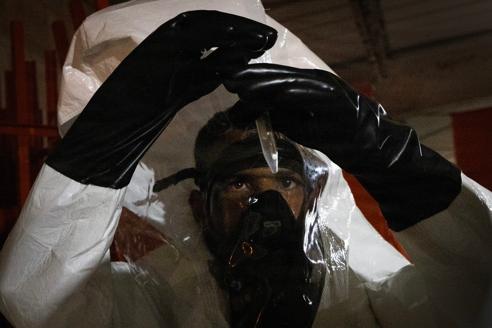 A person wearing full hazmat/protective gear including a white Tyvek suit, black rubber gloves, and a black respirator/gas mask is photographed in a dimly lit industrial or warehouse setting. The individual appears to be holding or manipulating a small object, possibly a vial or dropper, above their head. The image is shot through what appears to be a plastic barrier or the suit's hood, creating a layered visual effect.