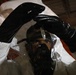 A person wearing full hazmat/protective gear including a white Tyvek suit, black rubber gloves, and a black respirator/gas mask is photographed in a dimly lit industrial or warehouse setting. The individual appears to be holding or manipulating a small object, possibly a vial or dropper, above their head. The image is shot through what appears to be a plastic barrier or the suit's hood, creating a layered visual effect.