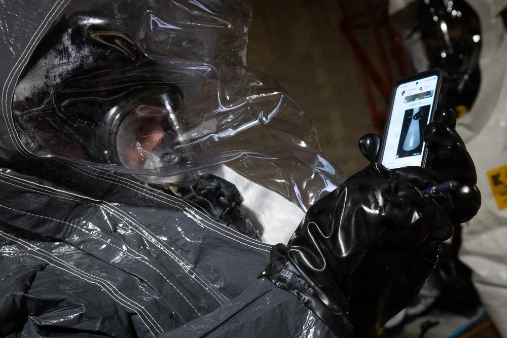 A person wearing a full hazmat/CBRN protective suit with a clear face shield and black rubber gloves is holding and viewing a smartphone. The suit appears to be a dark gray encapsulated chemical protection suit. A second similarly suited individual is partially visible in the background.