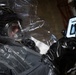 A person wearing a full hazmat/CBRN protective suit with a clear face shield and black rubber gloves is holding and viewing a smartphone. The suit appears to be a dark gray encapsulated chemical protection suit. A second similarly suited individual is partially visible in the background.