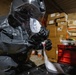 A person wearing a full black hazmat/protective suit with a clear face shield and black rubber gloves is handling what appears to be a test strip or sample collection material. The setting appears to be a warehouse or storage facility with stacked cardboard boxes visible in the background. The scene suggests a hazardous materials inspection, forensic investigation, or chemical detection operation.