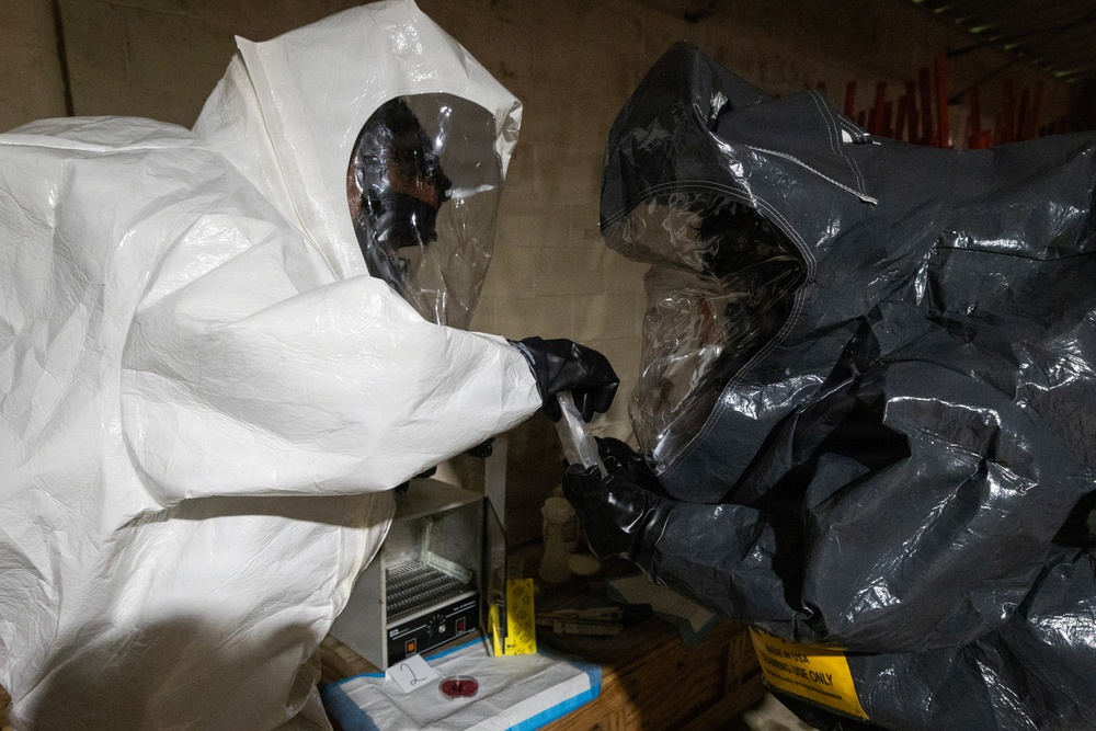 Two individuals wearing full hazmat/CBRN protective suits are engaged in what appears to be a sample handling or testing procedure. One person wears a white Tyvek-style suit and the other wears a dark gray/black encapsulated suit, both with full-face respirator visors and black gloves. They appear to be working with a small vial or sample container in an indoor setting with equipment visible in the background.