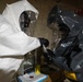 Two individuals wearing full hazmat/CBRN protective suits are engaged in what appears to be a sample handling or testing procedure. One person wears a white Tyvek-style suit and the other wears a dark gray/black encapsulated suit, both with full-face respirator visors and black gloves. They appear to be working with a small vial or sample container in an indoor setting with equipment visible in the background.