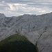 A dramatic alpine landscape featuring rugged, light-grey limestone mountain ridges with jagged rocky peaks. In the foreground, a lower green hill covered with coniferous forest contrasts sharply with the bare rocky slopes behind it. The scene is photographed under a partly cloudy sky with diffused natural light.