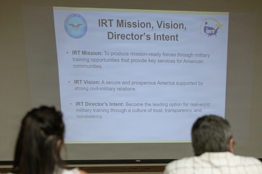 A projected presentation slide titled 'IRT Mission, Vision, Director's Intent' is displayed in what appears to be a briefing or classroom setting. Two individuals are seated in the foreground with their backs to the camera, viewing the slide. The slide contains three bullet points outlining the IRT (Innovative Readiness Training) program's mission, vision, and director's intent.