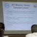 A projected presentation slide titled 'IRT Mission, Vision, Director's Intent' is displayed in what appears to be a briefing or classroom setting. Two individuals are seated in the foreground with their backs to the camera, viewing the slide. The slide contains three bullet points outlining the IRT (Innovative Readiness Training) program's mission, vision, and director's intent.