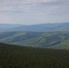 An elevated aerial or high-vantage-point view of a vast boreal forest landscape with rolling hills and mountains receding into the distance. Dense coniferous trees dominate the foreground, transitioning to mixed forest cover across multiple ridgelines. A faint road or trail is visible in the mid-ground, along with what appears to be a small structure or clearing on the right side.