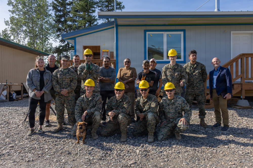 A group of approximately 16 people pose for a group photo in front of a newly constructed or renovated single-story building with blue trim. The group includes military personnel in camouflage uniforms, several wearing yellow hard hats, along with civilians. A small dog is visible in the foreground among the kneeling front row.