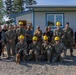 A group of approximately 16 people pose for a group photo in front of a newly constructed or renovated single-story building with blue trim. The group includes military personnel in camouflage uniforms, several wearing yellow hard hats, along with civilians. A small dog is visible in the foreground among the kneeling front row.