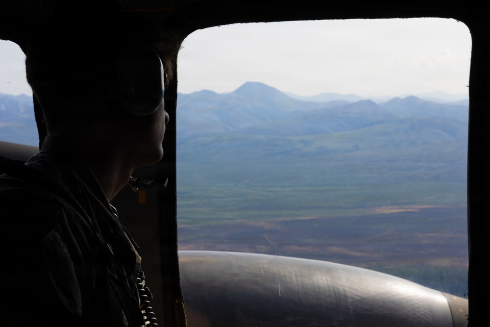 A military personnel member is seated inside an aircraft, viewed in silhouette, looking out through a large open window or door at a mountainous landscape below. The scene appears to be taken during an aerial mission over a remote, rugged terrain with rolling hills and valleys. A cylindrical metallic object, likely part of the aircraft's exterior or a fuel tank, is visible in the lower portion of the frame.